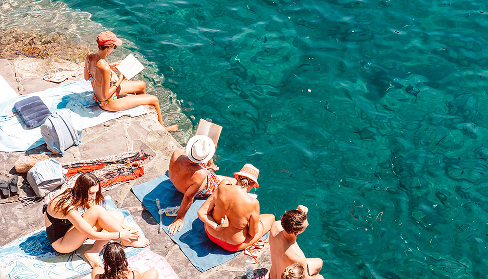 A summer scene in the Mediterranean. Locals and tourists sitting with their feet in the near perfect sea water, reading and relaxing in the sun.